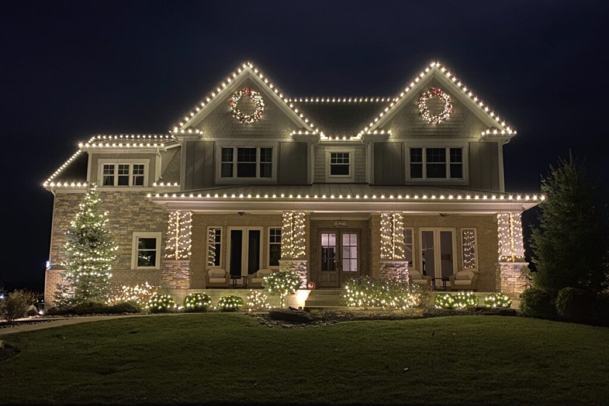 A two-story home with its entire roofline outlined in warm-white lights, wreaths on the gables and a lit evergreen in the yard.