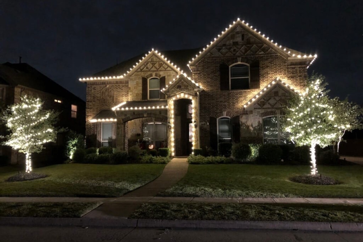 A brick home with a warm-white roofline and two front-yard trees wrapped trunk-to-branch in warm-white lights.