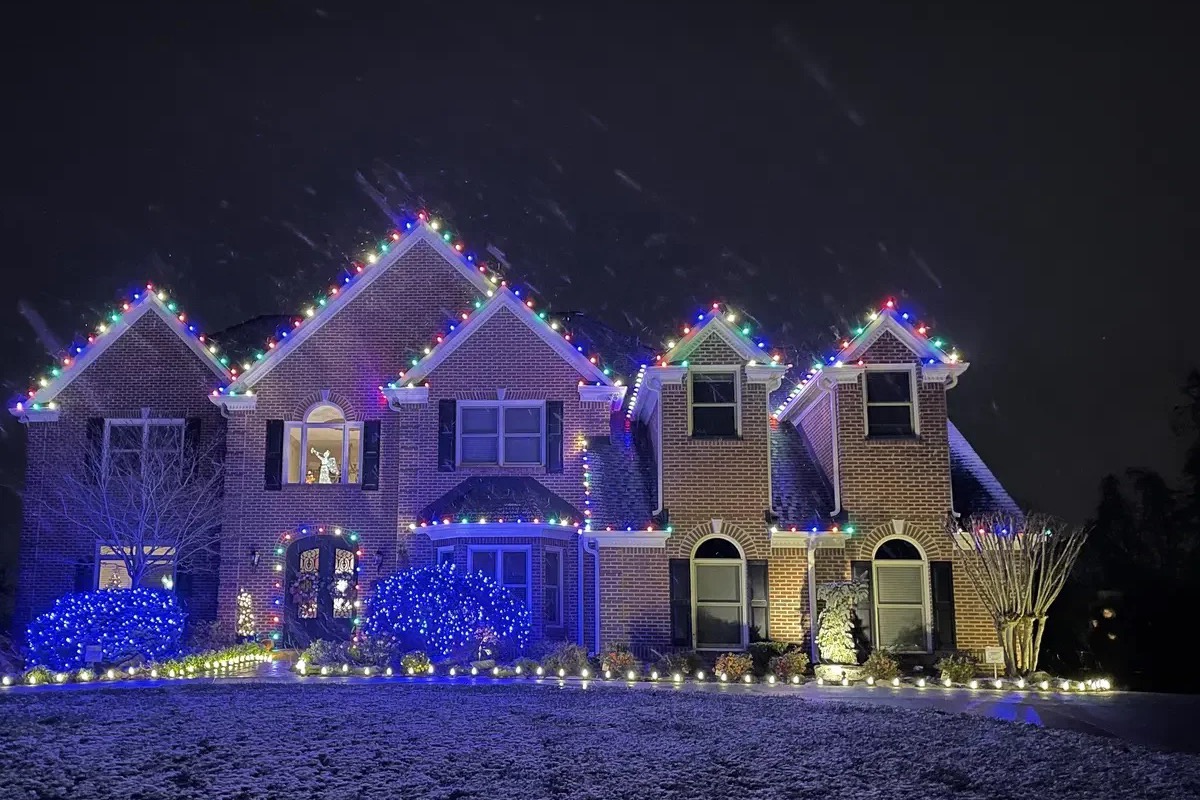 A brick home on a snowy night with a multicolor roofline and blue-lit bushes across the front yard.