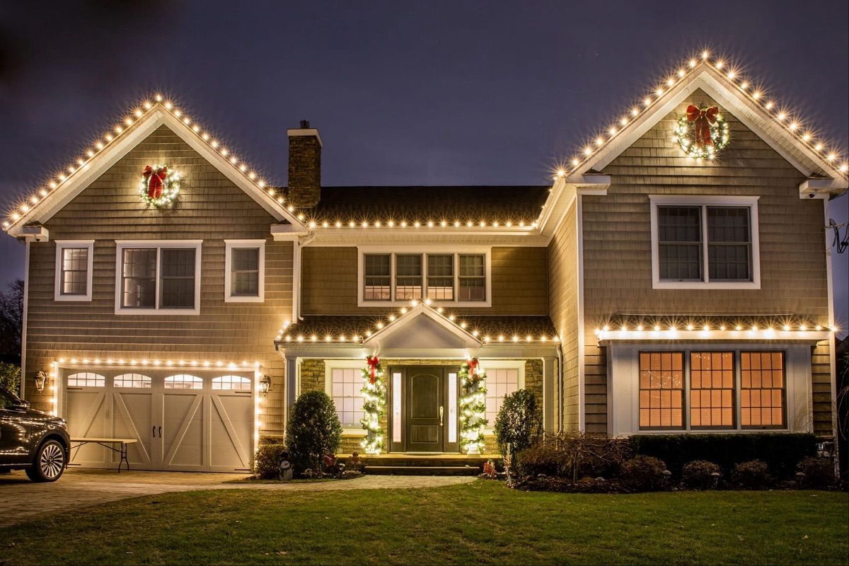 A large two-story home lit end to end with warm-white roofline lights, gable wreaths and wrapped entry columns.